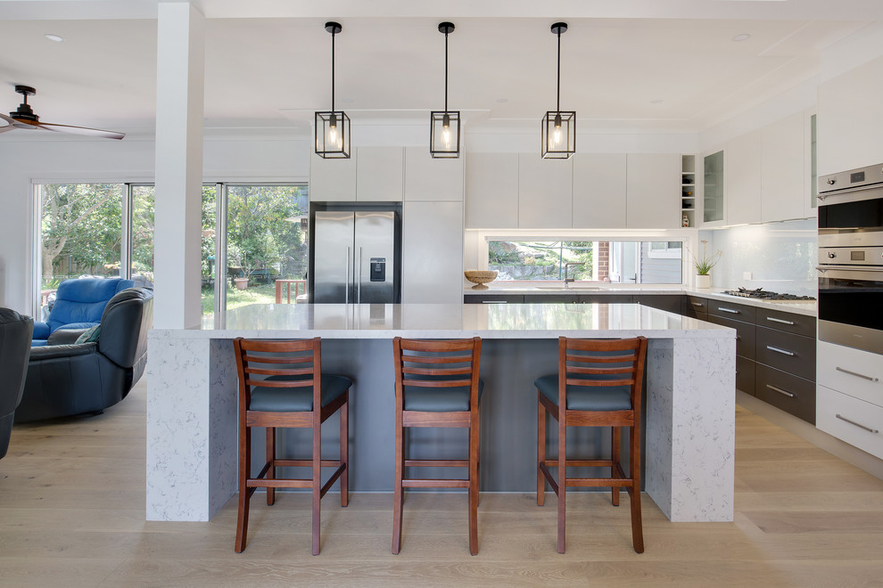 Bild på ett stort funkis grå grått kök, med en undermonterad diskho, släta luckor, vita skåp, bänkskiva i kvarts, vitt stänkskydd, glaspanel som stänkskydd, rostfria vitvaror, ljust trägolv och en köksö
