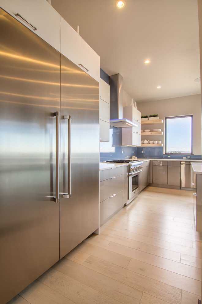 Photo of an expansive modern u-shaped open plan kitchen in Other with a belfast sink, flat-panel cabinets, grey cabinets, engineered stone countertops, blue splashback, glass sheet splashback, stainless steel appliances, light hardwood flooring, an island and beige floors.