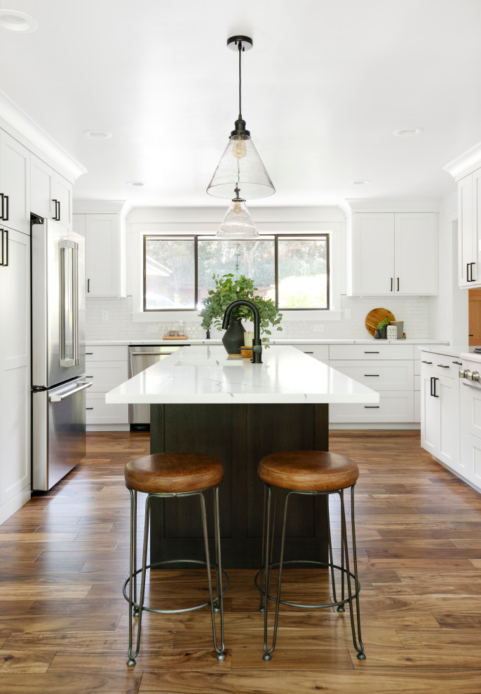 Inredning av ett lantligt mellanstort vit vitt kök, med en undermonterad diskho, skåp i shakerstil, vita skåp, bänkskiva i kvarts, vitt stänkskydd, stänkskydd i keramik, rostfria vitvaror, mellanmörkt trägolv, en köksö och brunt golv