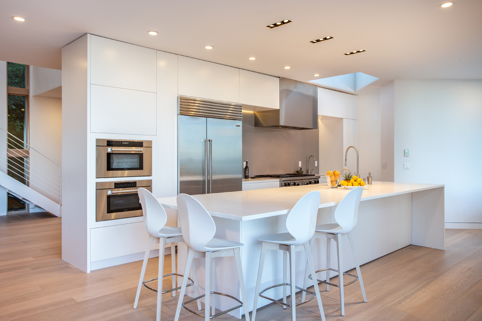 This is an example of a contemporary single-wall kitchen in Portland with a submerged sink, flat-panel cabinets, white cabinets, grey splashback, stainless steel appliances, medium hardwood flooring, an island, brown floors and white worktops.