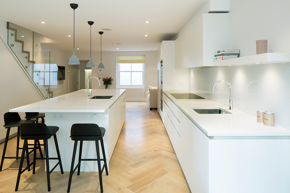 Example of a large minimalist single-wall light wood floor open concept kitchen design in London with a drop-in sink, flat-panel cabinets, white cabinets, quartzite countertops, gray backsplash, glass sheet backsplash, stainless steel appliances and an island