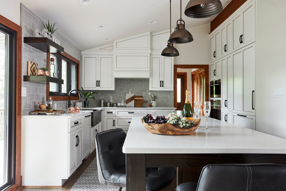 Cette image montre une cuisine américaine encastrable chalet en U de taille moyenne avec un évier de ferme, des portes de placard blanches, un plan de travail en verre, une crédence grise, une crédence en carreau de verre, un sol en bois brun, îlot, un sol marron, un plan de travail blanc et un placard à porte affleurante.