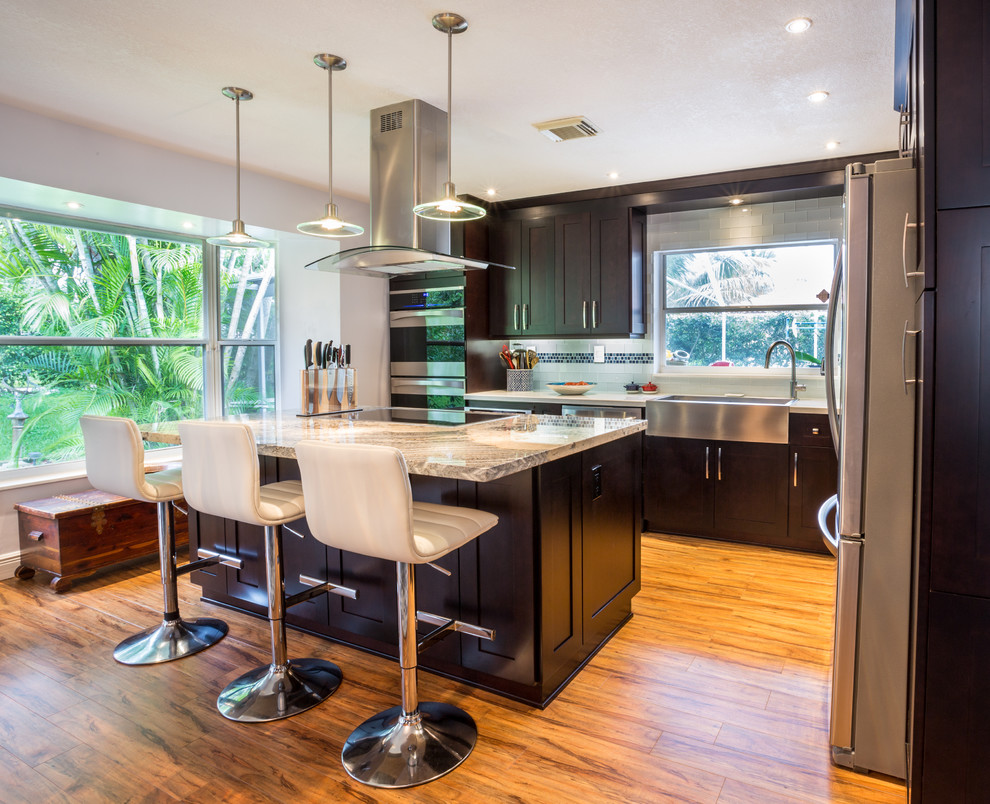 Large contemporary l-shaped kitchen/diner in Miami with a belfast sink, shaker cabinets, dark wood cabinets, granite worktops, multi-coloured splashback, glass tiled splashback, stainless steel appliances, medium hardwood flooring and an island.