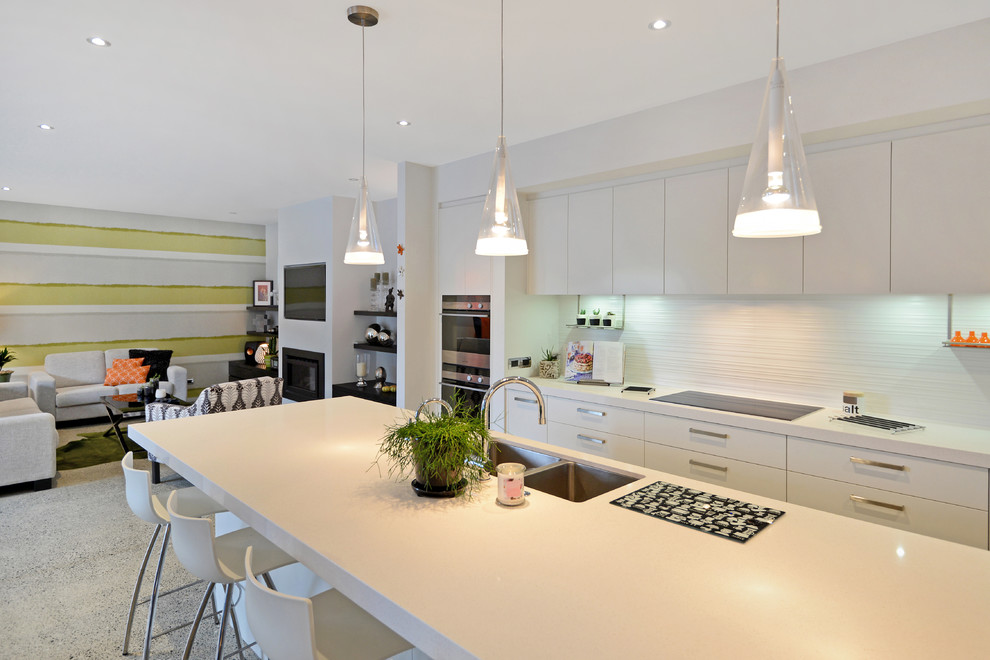 This is an example of a medium sized contemporary single-wall open plan kitchen in Auckland with a double-bowl sink, flat-panel cabinets, white cabinets, engineered stone countertops, white splashback, cement tile splashback, stainless steel appliances, concrete flooring and an island.