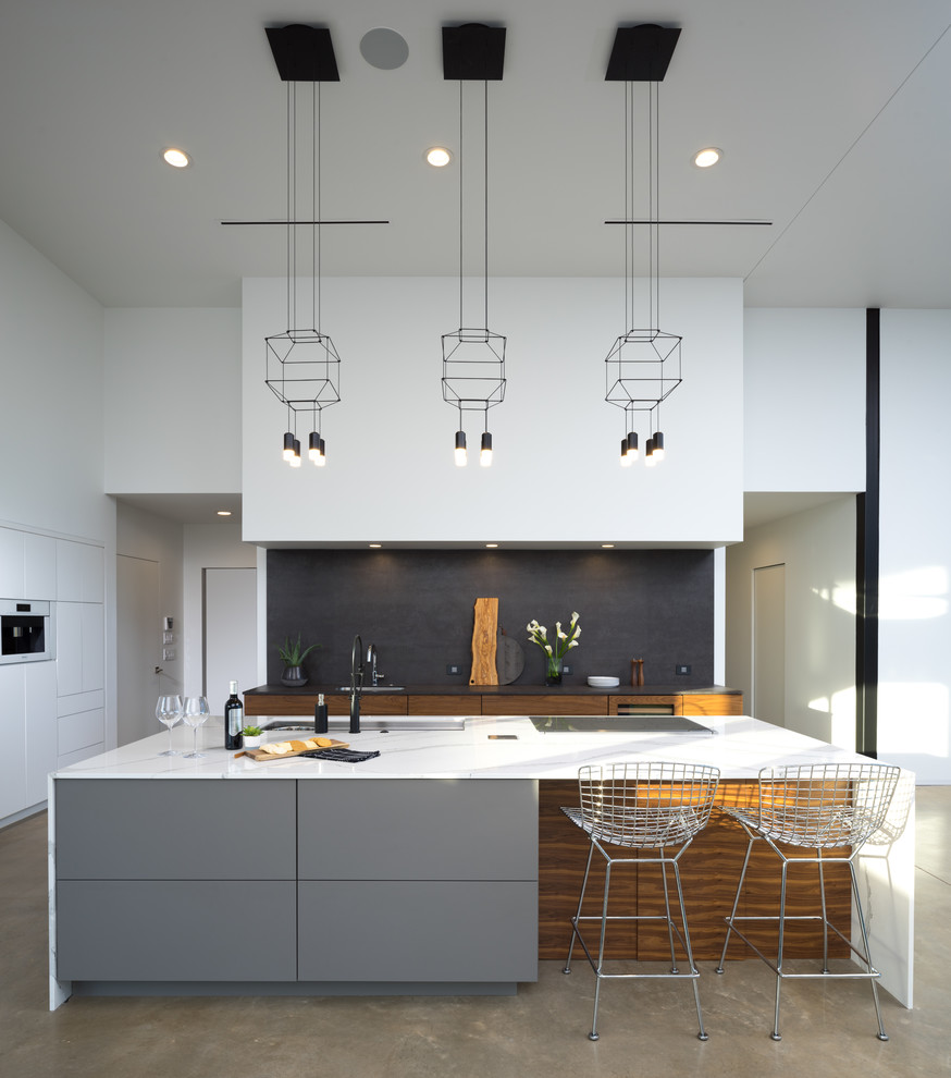 Стильный дизайн: угловая кухня-гостиная среднего размера в стиле модернизм с врезной мойкой, плоскими фасадами, столешницей из кварцевого агломерата, черным фартуком, белой техникой, бетонным полом и островом - последний тренд