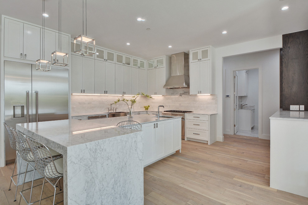 Photo of a large classic l-shaped open plan kitchen in Nashville with a belfast sink, shaker cabinets, white cabinets, marble worktops, white splashback, stainless steel appliances, light hardwood flooring and an island.
