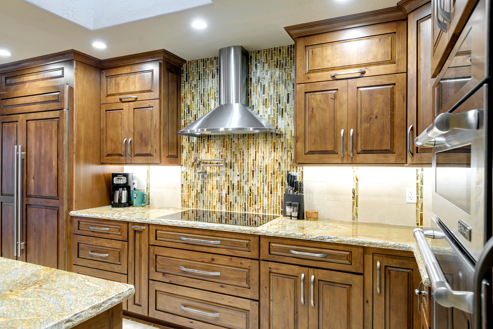 Example of a mid-sized mountain style l-shaped travertine floor open concept kitchen design in Phoenix with a drop-in sink, raised-panel cabinets, medium tone wood cabinets, granite countertops, green backsplash, mosaic tile backsplash, paneled appliances and an island