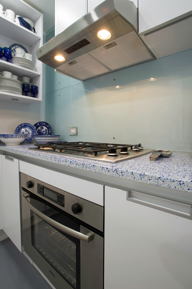 Foto på ett avskilt, litet funkis u-kök, med en undermonterad diskho, släta luckor, vita skåp, bänkskiva i återvunnet glas, blått stänkskydd, glaspanel som stänkskydd, rostfria vitvaror, betonggolv och en halv köksö