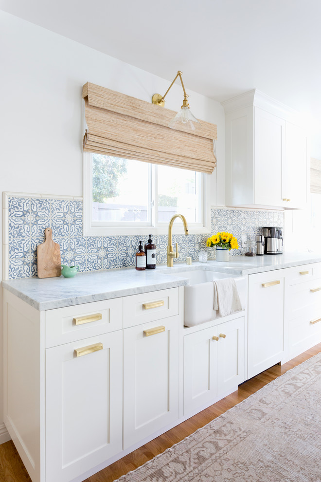 Modelo de cocina costera abierta con fregadero sobremueble, armarios estilo shaker, puertas de armario blancas, encimera de mármol, salpicadero azul, suelo de madera clara y suelo beige