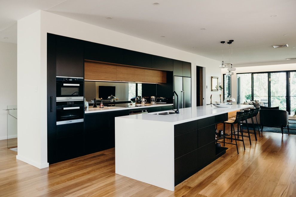 Cette image montre une grande cuisine parallèle minimaliste avec un évier encastré, des portes de placard noires, un plan de travail en quartz modifié, un électroménager noir, îlot et un plan de travail blanc.
