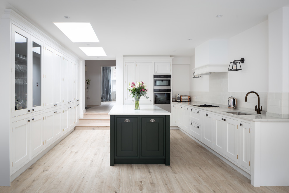 Large traditional l-shaped enclosed kitchen in London with a built-in sink, shaker cabinets, marble worktops, white splashback, marble splashback, stainless steel appliances, light hardwood flooring, an island, beige floors and white worktops.
