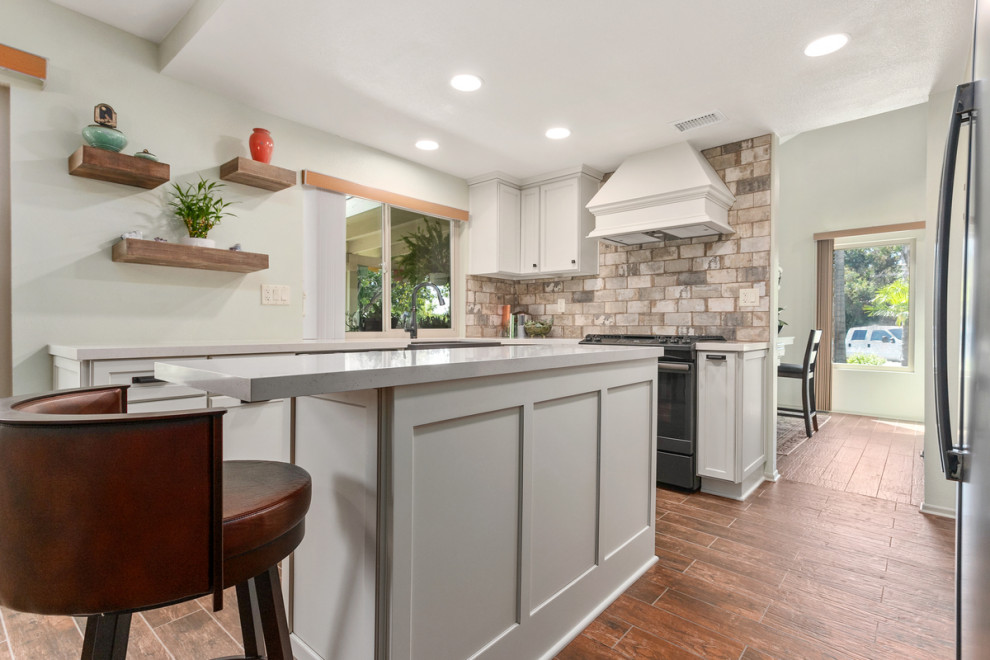 Small rustic l-shaped kitchen/diner in San Diego with a belfast sink, shaker cabinets, white cabinets, composite countertops, multi-coloured splashback, brick splashback, stainless steel appliances, ceramic flooring, an island, brown floors and white worktops.