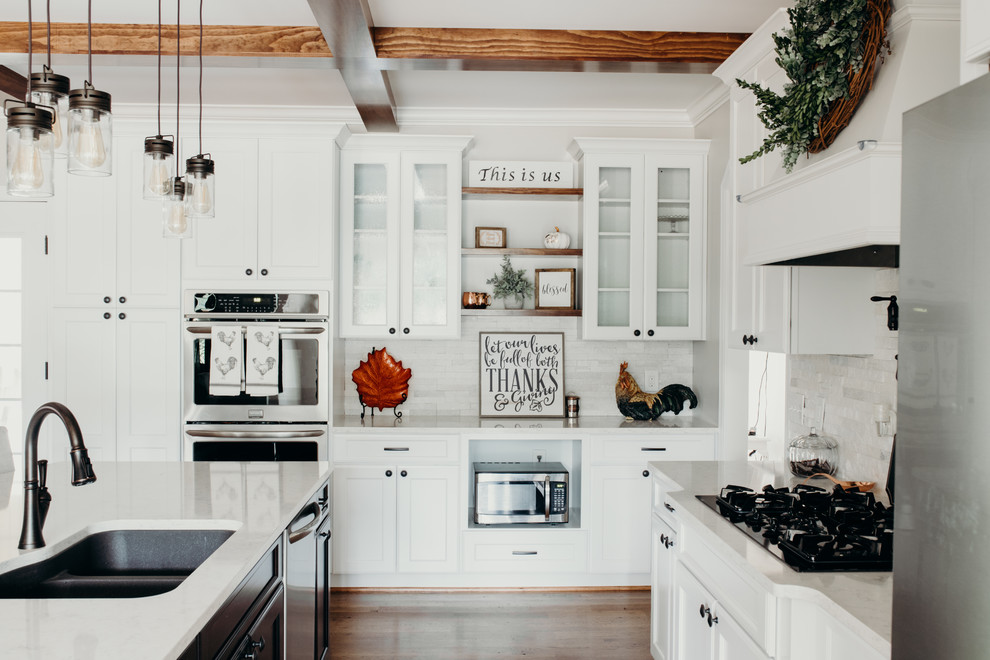 Inredning av ett lantligt stort vit vitt kök, med en undermonterad diskho, luckor med infälld panel, vita skåp, bänkskiva i kvarts, vitt stänkskydd, stänkskydd i stenkakel, rostfria vitvaror, mellanmörkt trägolv, en köksö och brunt golv