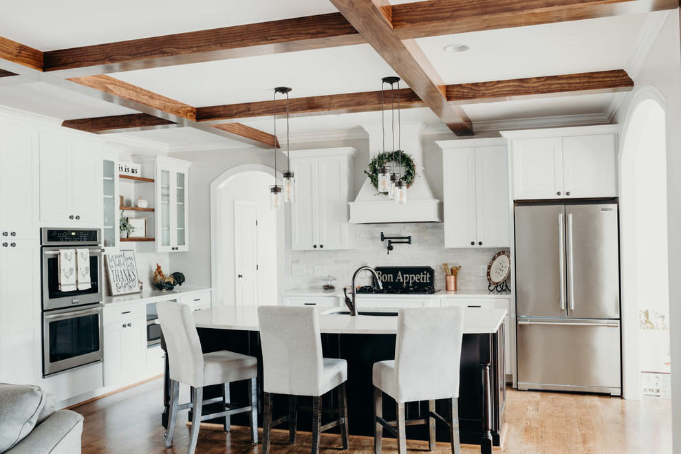 Inredning av ett lantligt stort vit vitt kök, med en undermonterad diskho, luckor med infälld panel, vita skåp, bänkskiva i kvarts, vitt stänkskydd, stänkskydd i stenkakel, rostfria vitvaror, mellanmörkt trägolv, en köksö och brunt golv