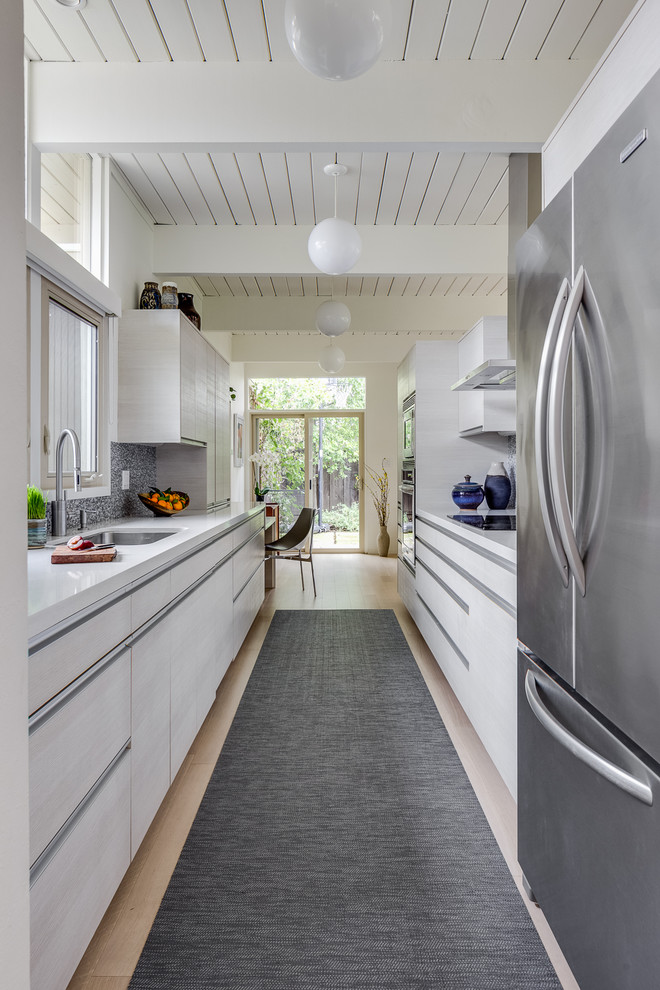 Example of a mid-sized minimalist galley light wood floor, beige floor and shiplap ceiling enclosed kitchen design in San Francisco with flat-panel cabinets, white cabinets, solid surface countertops, mosaic tile backsplash, stainless steel appliances, an undermount sink, white countertops and gray backsplash