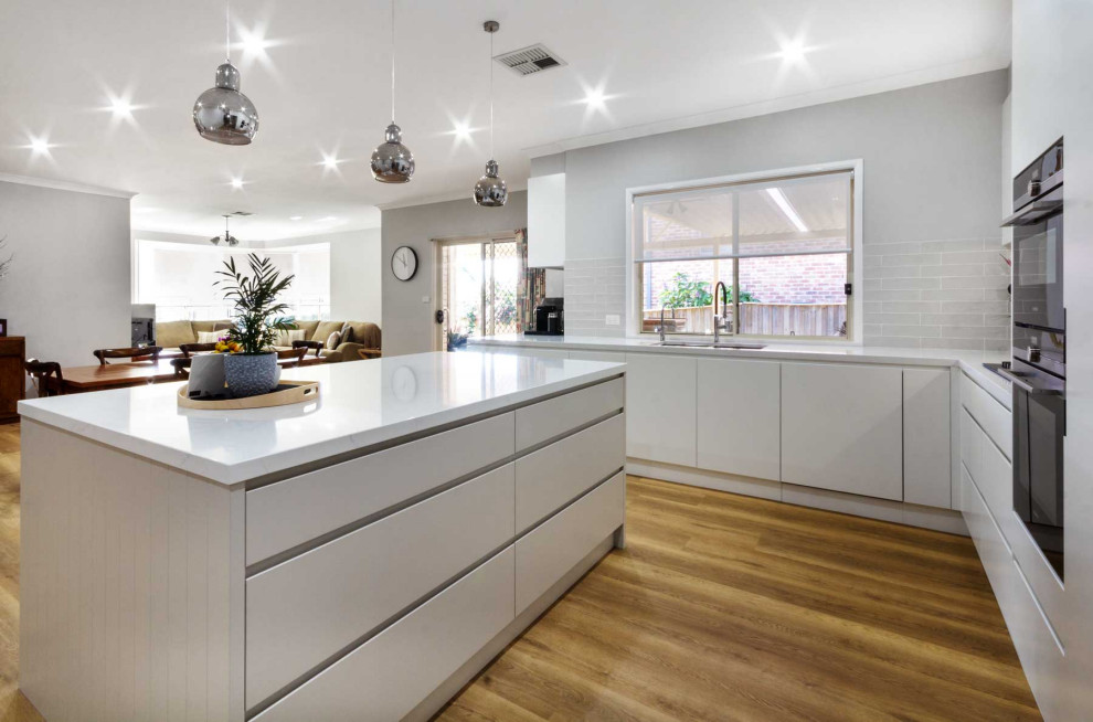 This is an example of a medium sized contemporary l-shaped open plan kitchen in Sydney with a double-bowl sink, flat-panel cabinets, white cabinets, engineered stone countertops, grey splashback, ceramic splashback, black appliances, vinyl flooring, an island, brown floors and white worktops.