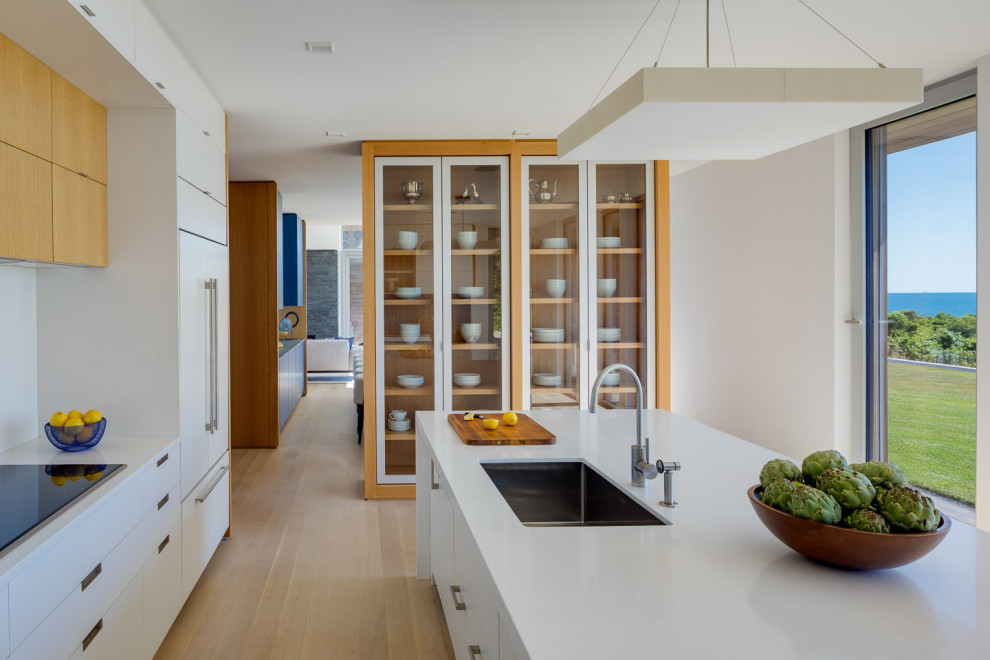 Photo of a large contemporary l-shaped open plan kitchen in Boston with a submerged sink, flat-panel cabinets, white cabinets, quartz worktops, blue splashback, glass sheet splashback, integrated appliances, light hardwood flooring, an island, beige floors and white worktops.