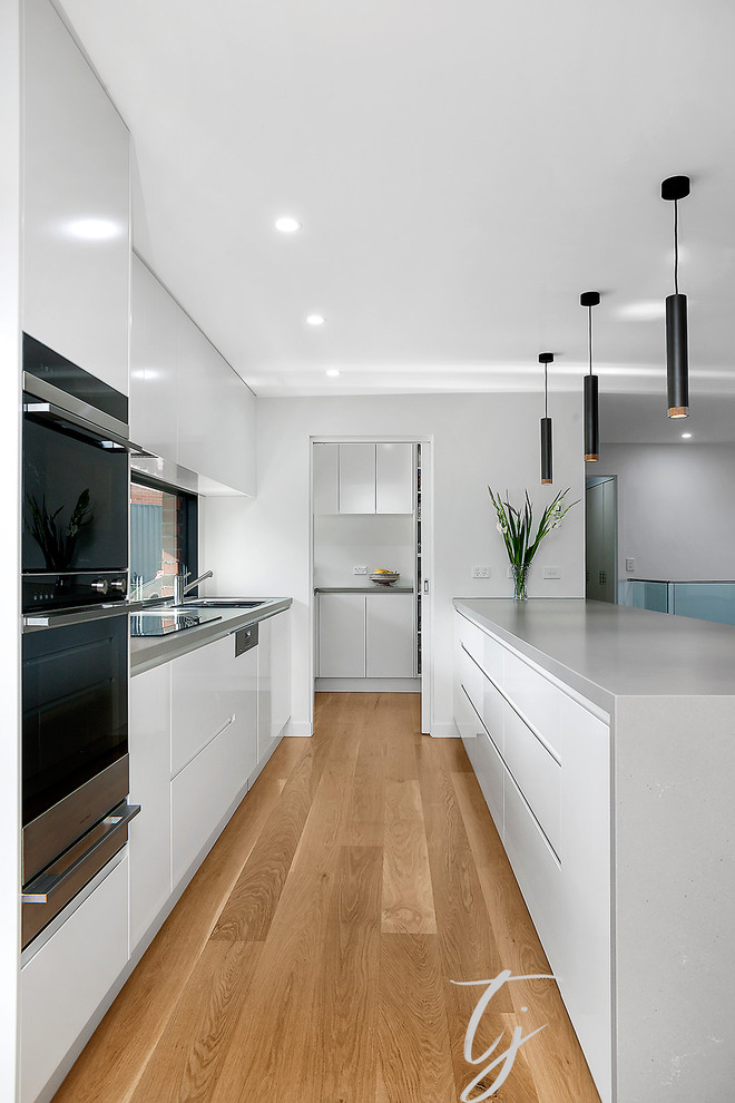 Design ideas for a large contemporary galley kitchen pantry in Melbourne with a double-bowl sink, white cabinets, composite countertops, wood splashback, stainless steel appliances, light hardwood flooring, an island and white worktops.