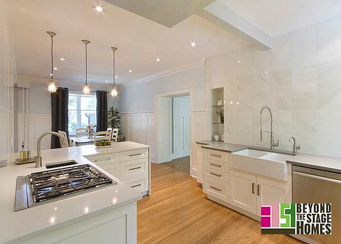 This is an example of a classic kitchen/diner in Other with a belfast sink, shaker cabinets, white cabinets, engineered stone countertops, white splashback, stone tiled splashback, stainless steel appliances, light hardwood flooring and a breakfast bar.