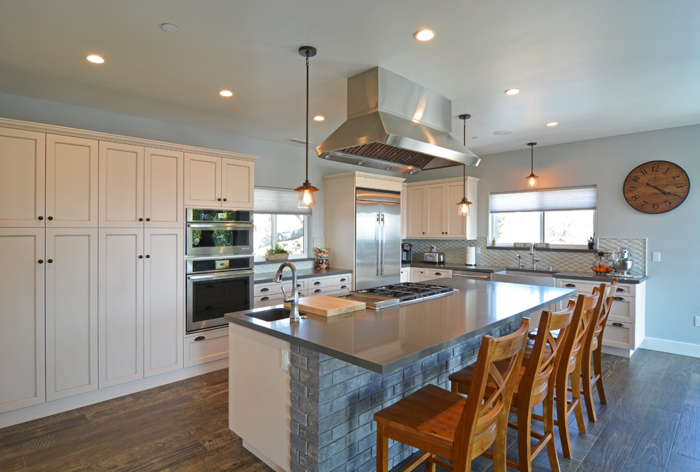 This is an example of a medium sized traditional l-shaped kitchen/diner in San Luis Obispo with a belfast sink, recessed-panel cabinets, white cabinets, engineered stone countertops, white splashback, ceramic splashback, stainless steel appliances, ceramic flooring, an island, brown floors and grey worktops.