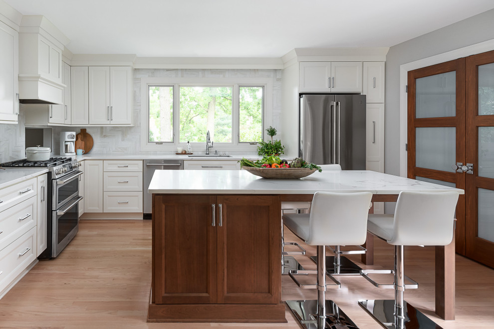 Photo of a medium sized traditional l-shaped open plan kitchen in Chicago with a submerged sink, flat-panel cabinets, white cabinets, engineered stone countertops, white splashback, marble splashback, stainless steel appliances, light hardwood flooring, an island, brown floors and white worktops.