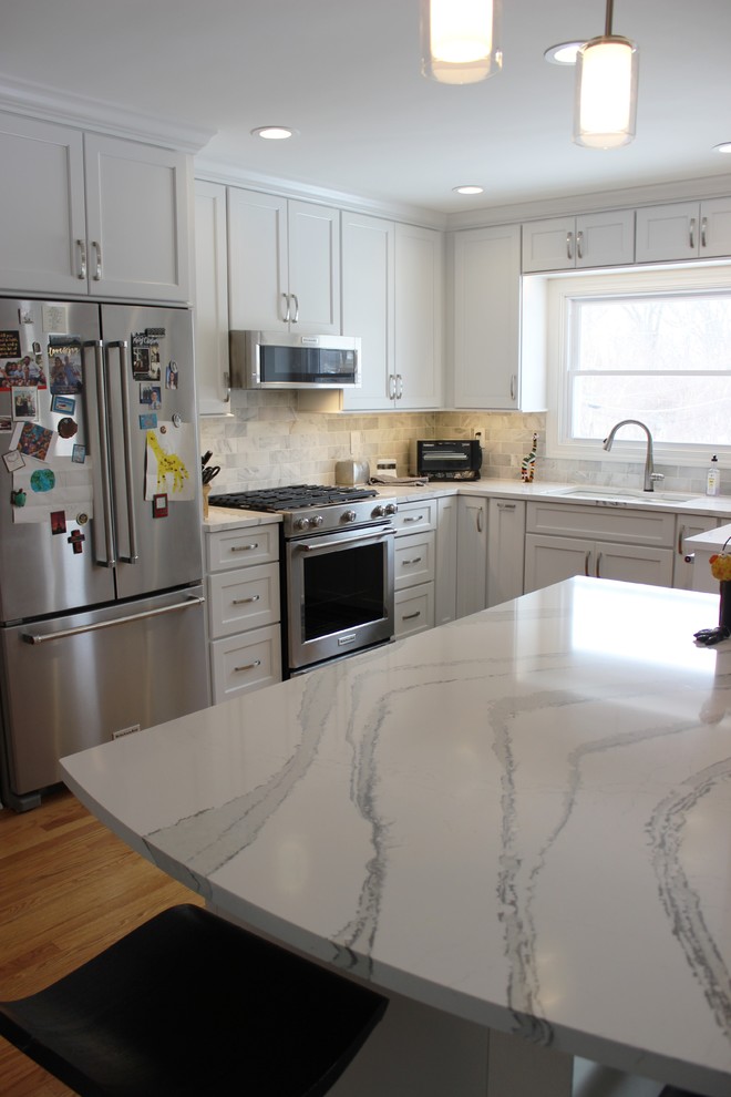 Klassisk inredning av ett mellanstort vit vitt u-kök, med en undermonterad diskho, släta luckor, vita skåp, bänkskiva i kvarts, grått stänkskydd, stänkskydd i marmor, rostfria vitvaror, ljust trägolv och en halv köksö