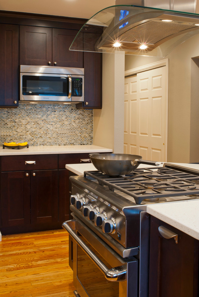Inredning av ett modernt litet kök, med luckor med upphöjd panel, bruna skåp, bänkskiva i kvarts, flerfärgad stänkskydd, stänkskydd i glaskakel, rostfria vitvaror, ljust trägolv och en köksö