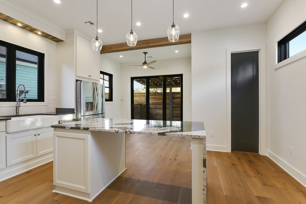 Inspiration for a small farmhouse l-shaped light wood floor and brown floor eat-in kitchen remodel in New Orleans with a farmhouse sink, flat-panel cabinets, white cabinets, granite countertops, white backsplash, subway tile backsplash, stainless steel appliances, an island and black countertops
