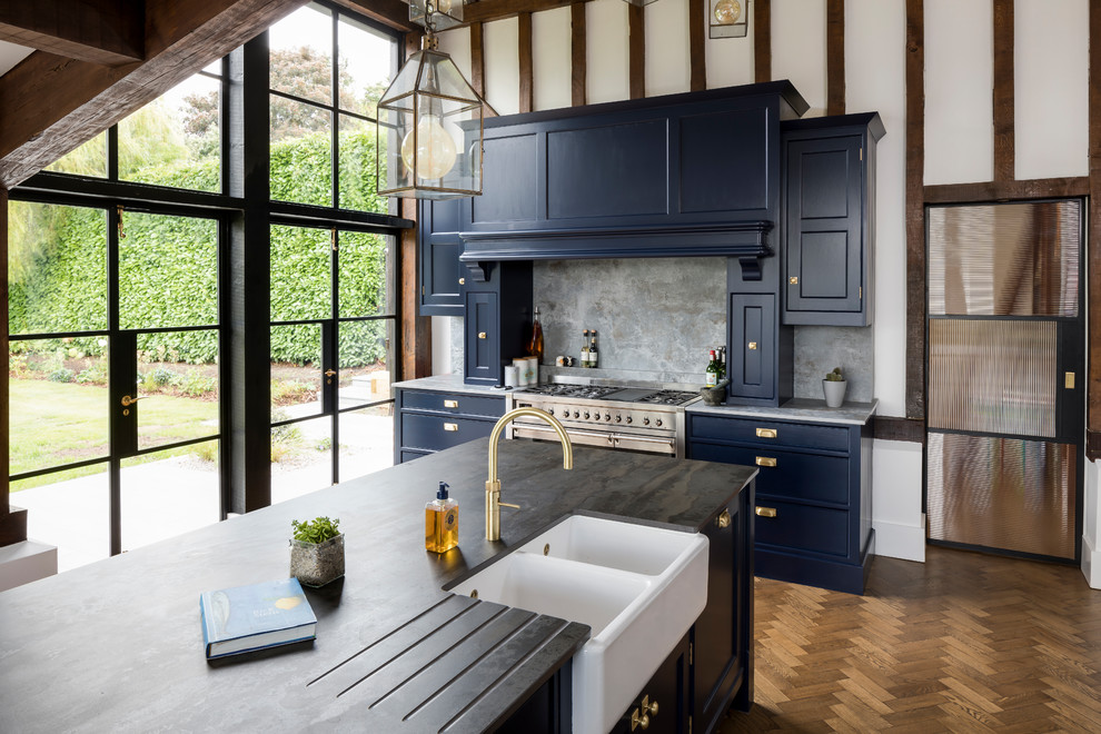 Medium sized farmhouse kitchen in Essex with a belfast sink, recessed-panel cabinets, blue cabinets, grey splashback, stainless steel appliances, an island, brown floors and black worktops.