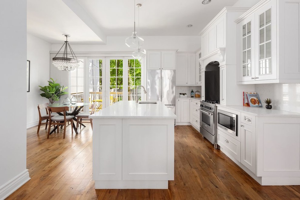 Design ideas for a traditional l-shaped kitchen/diner in New York with a submerged sink, white cabinets, white splashback, stainless steel appliances, medium hardwood flooring, an island, white worktops and recessed-panel cabinets.