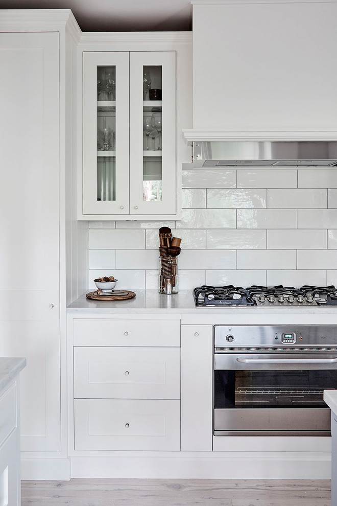 Photo of a medium sized classic u-shaped kitchen pantry in Sydney with a belfast sink, shaker cabinets, white cabinets, marble worktops, white splashback, metro tiled splashback, stainless steel appliances, light hardwood flooring and an island.
