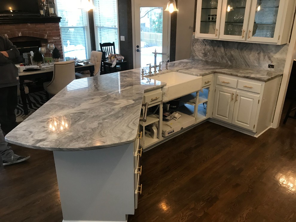 This is an example of a medium sized modern galley kitchen/diner in Kansas City with a belfast sink, raised-panel cabinets, white cabinets, marble worktops, white splashback, marble splashback, stainless steel appliances, medium hardwood flooring, a breakfast bar, brown floors and white worktops.