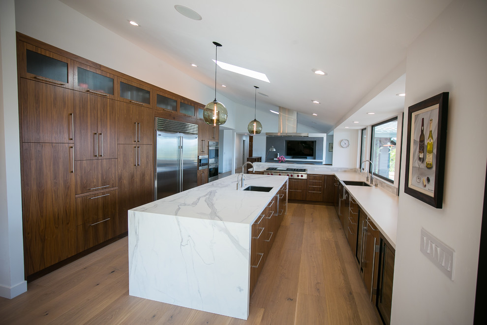 Example of a large trendy l-shaped light wood floor open concept kitchen design in San Francisco with a farmhouse sink, flat-panel cabinets, medium tone wood cabinets, solid surface countertops, white backsplash, porcelain backsplash, stainless steel appliances and an island