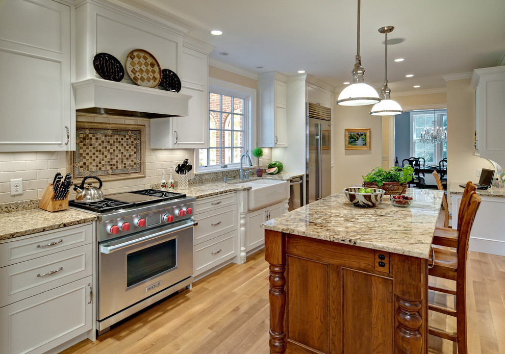 Inspiration for a large traditional galley kitchen in Minneapolis with a belfast sink, recessed-panel cabinets, white cabinets, granite worktops, stainless steel appliances, light hardwood flooring and an island.