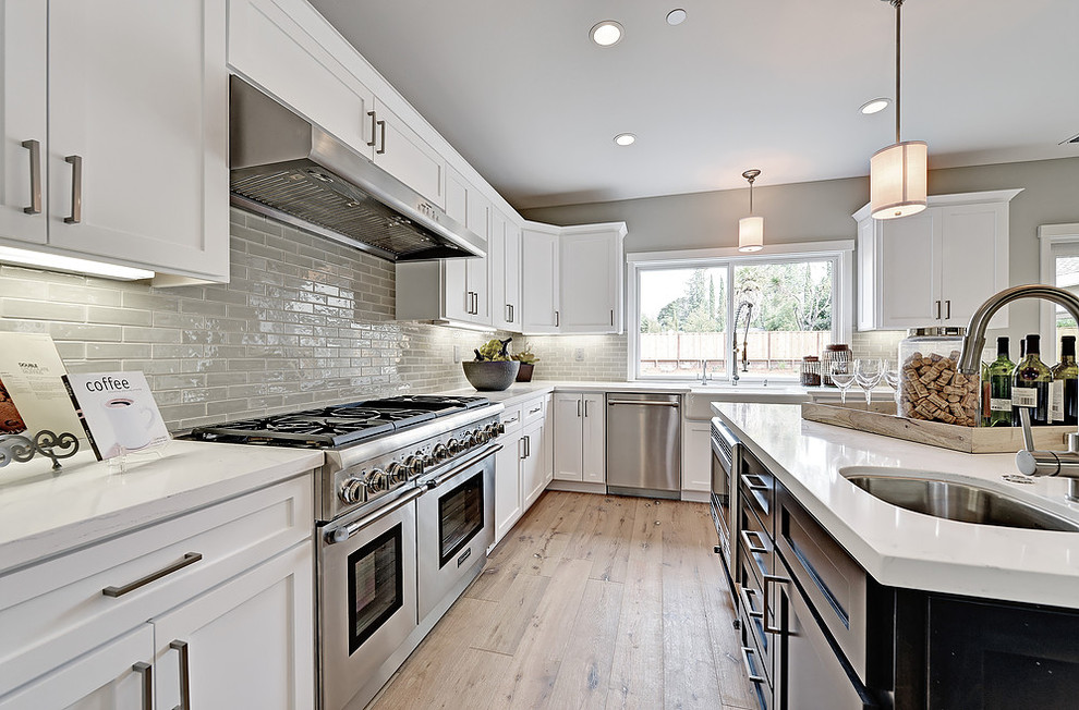 Transitional u-shaped light wood floor eat-in kitchen photo in San Francisco with a farmhouse sink, shaker cabinets, white cabinets, gray backsplash, glass tile backsplash, stainless steel appliances, an island and solid surface countertops
