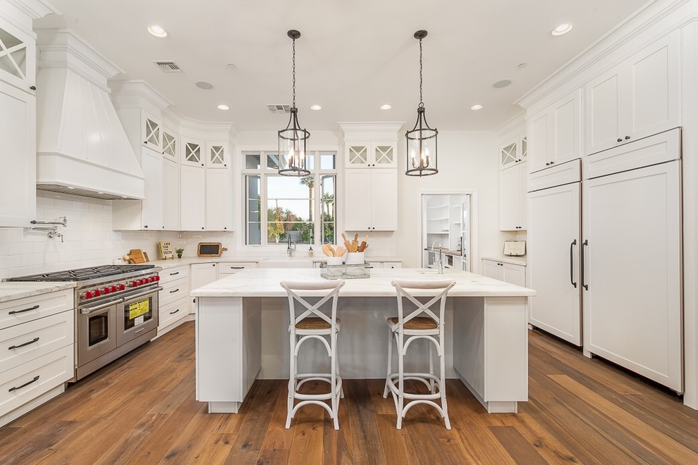 Design ideas for an expansive traditional u-shaped kitchen in Phoenix with a belfast sink, shaker cabinets, white cabinets, white splashback, metro tiled splashback, integrated appliances, medium hardwood flooring and an island.