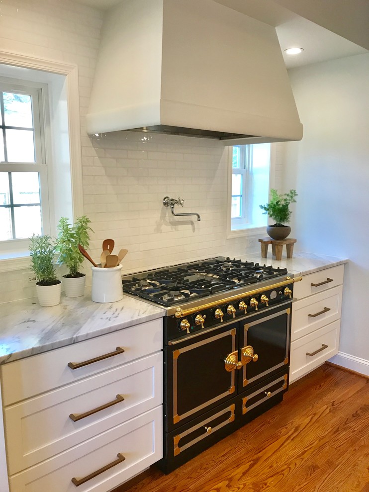 Example of a large farmhouse u-shaped medium tone wood floor and brown floor open concept kitchen design in DC Metro with a farmhouse sink, shaker cabinets, white cabinets, marble countertops, white backsplash, ceramic backsplash, black appliances and an island