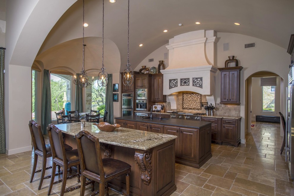 Large traditional l-shaped enclosed kitchen in Austin with a submerged sink, granite worktops, stainless steel appliances, travertine flooring, multiple islands, raised-panel cabinets, medium wood cabinets, beige splashback and brown floors.