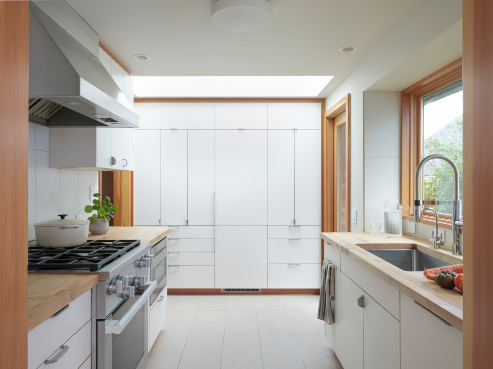 Example of a mid-sized minimalist galley ceramic tile and gray floor enclosed kitchen design with a single-bowl sink, flat-panel cabinets, white cabinets, wood countertops, beige backsplash, ceramic backsplash, stainless steel appliances and no island