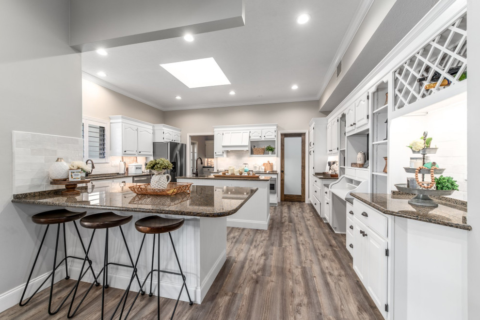 This is an example of a large farmhouse u-shaped kitchen pantry in Miami with a submerged sink, raised-panel cabinets, white cabinets, granite worktops, white splashback, metro tiled splashback, stainless steel appliances, vinyl flooring, an island, brown floors and brown worktops.