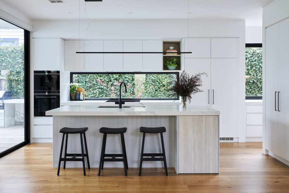 Foto på ett funkis vit kök med öppen planlösning, med en undermonterad diskho, släta luckor, vita skåp, fönster som stänkskydd, integrerade vitvaror, mellanmörkt trägolv, en köksö och brunt golv