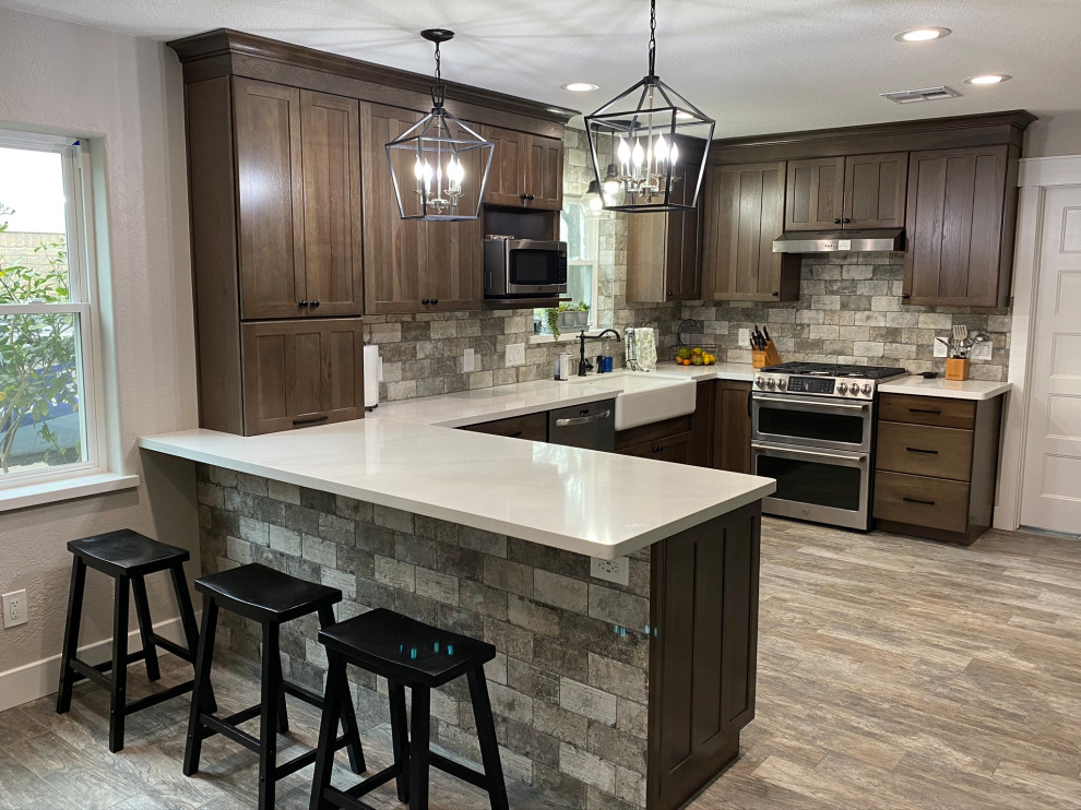 This is an example of a medium sized classic u-shaped kitchen/diner in Houston with a belfast sink, shaker cabinets, brown cabinets, wood worktops, grey splashback, travertine splashback, stainless steel appliances, porcelain flooring, a breakfast bar, grey floors and white worktops.