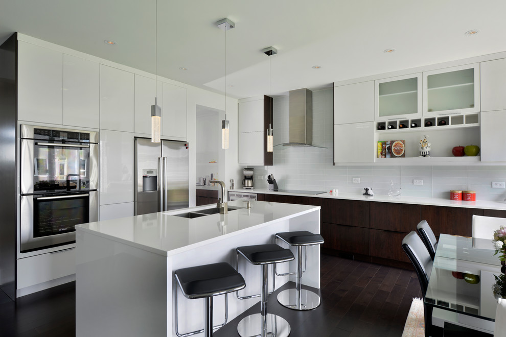 Photo of a contemporary l-shaped kitchen/diner in Ottawa with flat-panel cabinets, white cabinets, white splashback, a double-bowl sink, dark hardwood flooring, stainless steel appliances, an island and glass tiled splashback.
