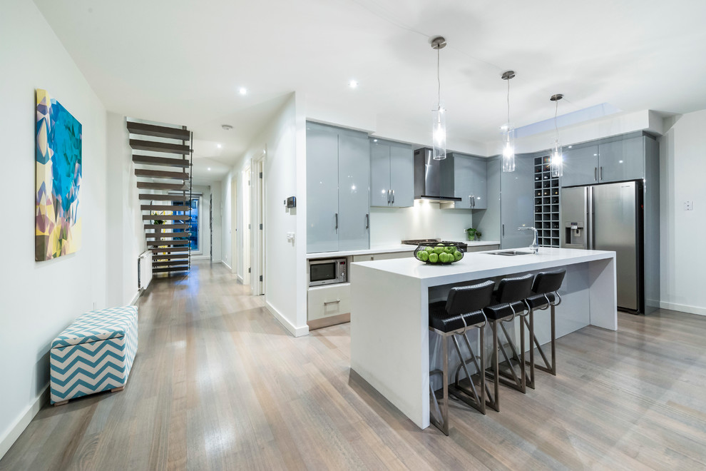 This is an example of a medium sized contemporary l-shaped kitchen/diner in Melbourne with a submerged sink, flat-panel cabinets, grey cabinets, engineered stone countertops, white splashback, glass sheet splashback, stainless steel appliances, light hardwood flooring and an island.