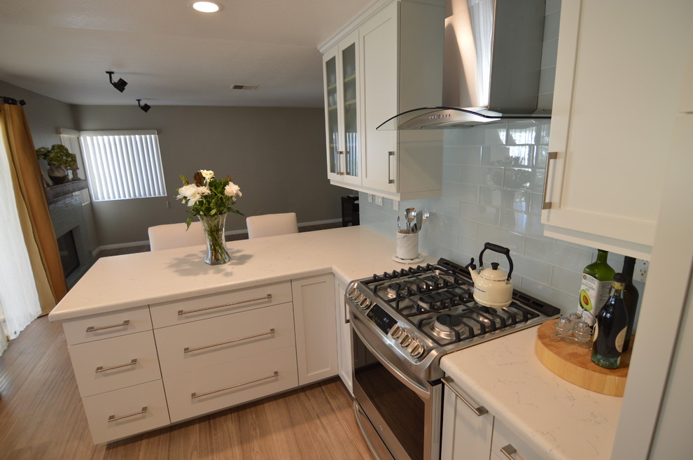 Medium sized modern open plan kitchen in Los Angeles with a belfast sink, recessed-panel cabinets, white cabinets, engineered stone countertops, blue splashback, glass tiled splashback, stainless steel appliances, laminate floors, no island, brown floors and white worktops.