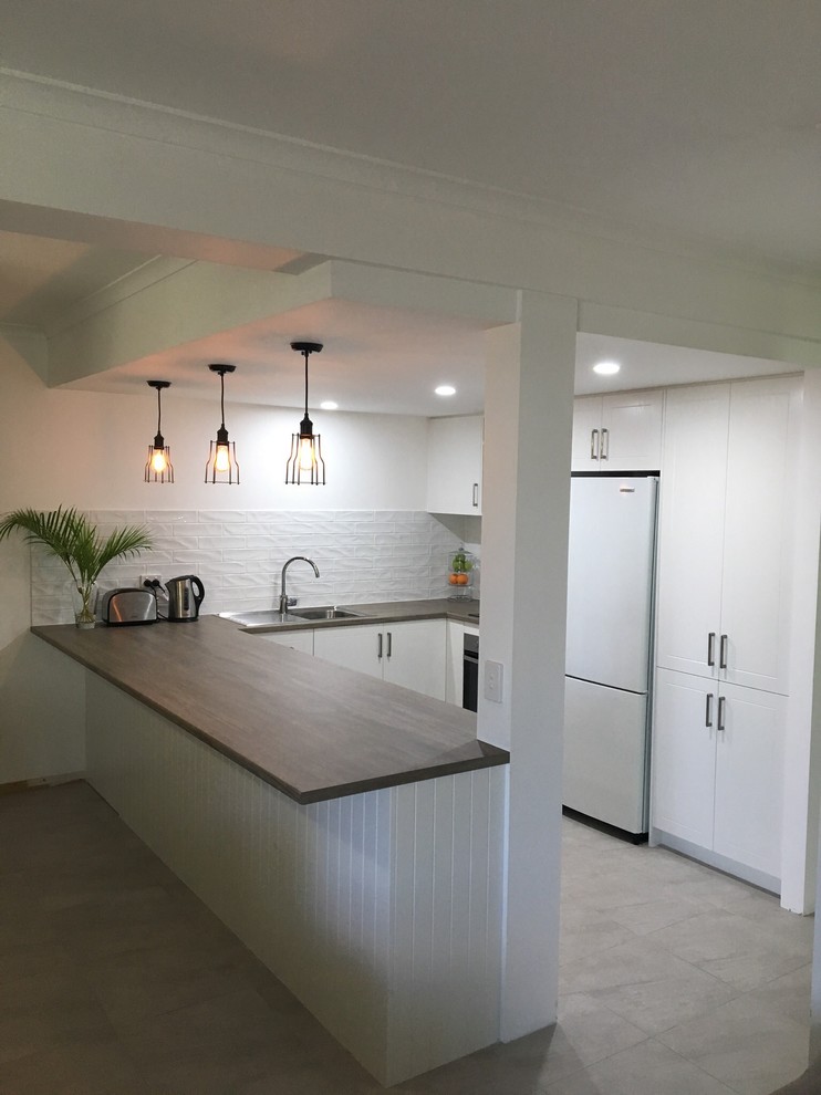 Photo of a small contemporary u-shaped open plan kitchen in Brisbane with a built-in sink, shaker cabinets, white cabinets, laminate countertops, white splashback, metro tiled splashback, stainless steel appliances, ceramic flooring, a breakfast bar and grey floors.