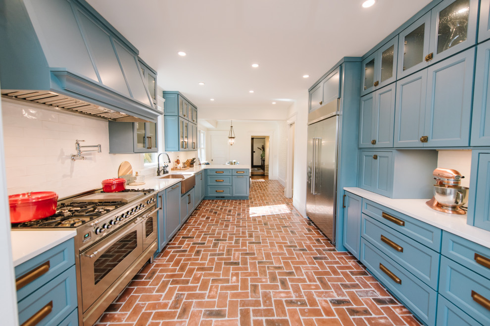 Photo of a medium sized bohemian galley kitchen/diner in Atlanta with a belfast sink, flat-panel cabinets, blue cabinets, engineered stone countertops, white splashback, porcelain splashback, stainless steel appliances, brick flooring, a breakfast bar and white worktops.