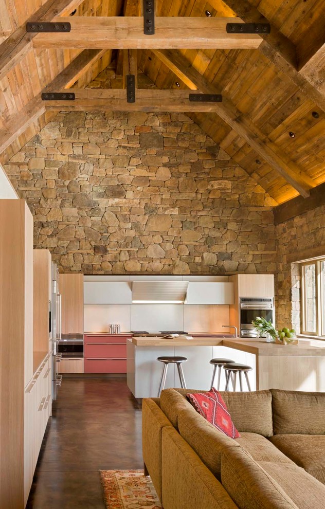 Imagen de cocina rural abierta con armarios con paneles lisos, electrodomésticos de acero inoxidable, puertas de armario de madera clara, salpicadero blanco y pared de piedra