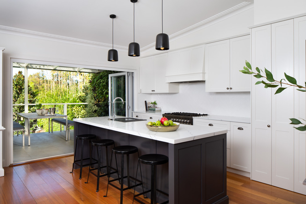 Exemple d'une cuisine parallèle chic avec un placard à porte shaker, des portes de placard blanches, une crédence blanche, un électroménager noir, un sol en bois brun, îlot, un sol marron et un plan de travail blanc.
