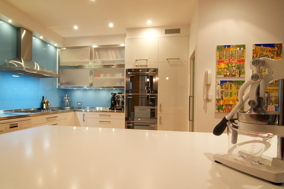 Idéer för ett avskilt, mellanstort modernt vit l-kök, med en integrerad diskho, släta luckor, vita skåp, bänkskiva i rostfritt stål, blått stänkskydd, glaspanel som stänkskydd, rostfria vitvaror, ljust trägolv och en köksö